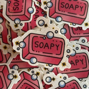 May include: A stack of pink and white stickers with a cartoon illustration of a bar of soap with the word "SOAPY" printed on it. The soap is surrounded by white bubbles.