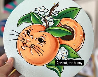 May include: A hand-painted round canvas with a peach-colored fruit with a cat face and whiskers. The fruit is surrounded by green leaves and white flowers. The text "Apricot, the bunny" is written at the bottom of the canvas.