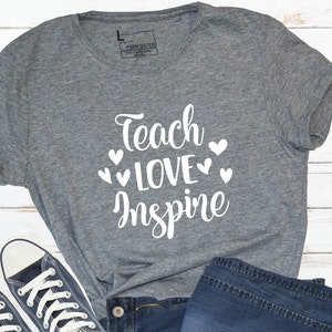 May include: A heather grey t-shirt with the text "Teach Love Inspire" in white lettering with small white hearts around the text.