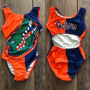 May include: Orange and blue leotard with a gator graphic and the word "Gators" on the front. The leotard has a cutout in the back.