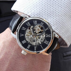 K&ouml;nnte beinhalten: Eine silberfarbene Armbanduhr mit schwarzem Zifferblatt, wei&szlig;en r&ouml;mischen Ziffern und einem sichtbaren skelettierten mechanischen Uhrwerk. Die Uhr hat ein schwarzes Lederarmband mit strukturierter Oberfl&auml;che und ein rundes Geh&auml;use aus poliertem Metall.