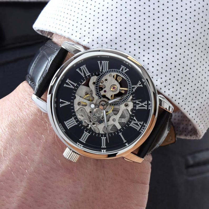 K&ouml;nnte beinhalten: Eine silberfarbene Armbanduhr mit schwarzem Zifferblatt, wei&szlig;en r&ouml;mischen Ziffern und einem sichtbaren skelettierten mechanischen Uhrwerk. Die Uhr hat ein schwarzes Lederarmband mit strukturierter Oberfl&auml;che und ein rundes Geh&auml;use aus poliertem Metall.