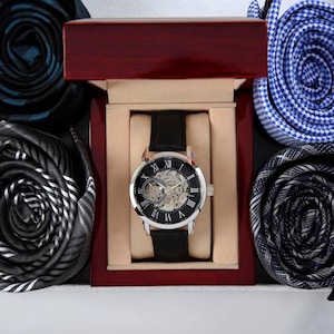 K&ouml;nnte beinhalten: Eine mechanische Armbanduhr mit schwarzem Lederarmband und silberfarbenem Geh&auml;use mit Skelett-Zifferblatt und r&ouml;mischen Ziffern, pr&auml;sentiert in einer polierten dunklen Holzschatulle, umgeben von vier gerollten Krawatten in verschiedenen Mustern.