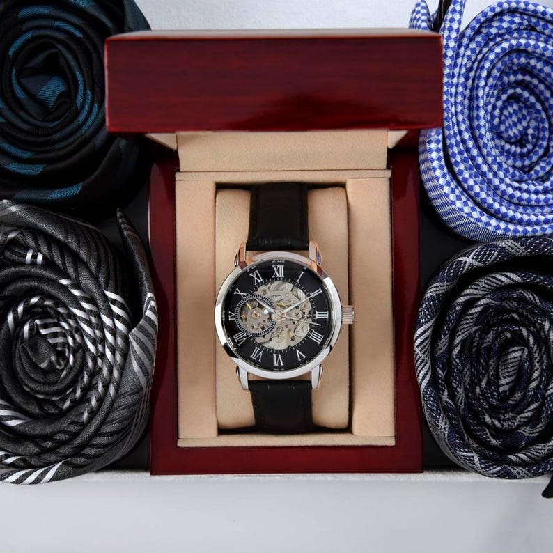 K&ouml;nnte beinhalten: Eine mechanische Armbanduhr mit schwarzem Lederarmband und silberfarbenem Geh&auml;use mit Skelett-Zifferblatt und r&ouml;mischen Ziffern, pr&auml;sentiert in einer polierten dunklen Holzschatulle, umgeben von vier gerollten Krawatten in verschiedenen Mustern.