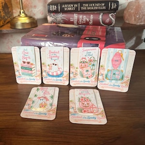 May include: Six magnetic bookmarks featuring pastel illustrations of ghosts, pumpkins, and beach items. Each bookmark is attached to a branded card with the text Booked at the Spooky Resort. Designs include a stack of books, a ghost in a pool float, and travel gear.