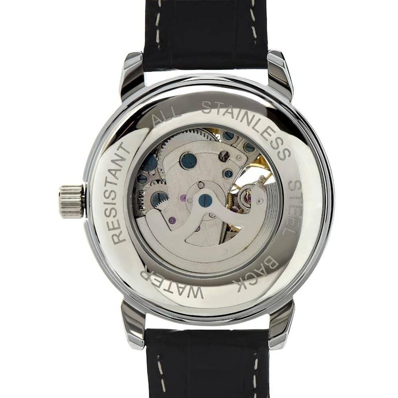 K&ouml;nnte beinhalten: Eine Armbanduhr mit schwarzem Lederarmband und silberfarbenem, kreisf&ouml;rmigem Geh&auml;use. Die Geh&auml;user&uuml;ckseite ist transparent und gibt den Blick auf das mechanische Uhrwerk frei. Der Metallrand tr&auml;gt die Gravur: ALL STAINLESS STEEL WATER RESISTANT.