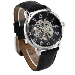 K&ouml;nnte beinhalten: Eine Armbanduhr mit einem runden silberfarbenen Geh&auml;use und einem schwarzen Zifferblatt mit wei&szlig;en r&ouml;mischen Ziffern. Die Mitte des Zifferblatts ist skelettiert und gibt den Blick auf das mechanische Uhrwerk frei. Mit schwarzem Lederarmband.