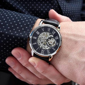 K&ouml;nnte beinhalten: Eine Armbanduhr mit rundem, silberfarbenem Geh&auml;use und schwarzem Zifferblatt mit wei&szlig;en r&ouml;mischen Ziffern. Die Mitte des Zifferblatts ist skelettiert und gibt den Blick auf das mechanische Uhrwerk frei. Die Uhr hat ein schwarzes Lederarmband.