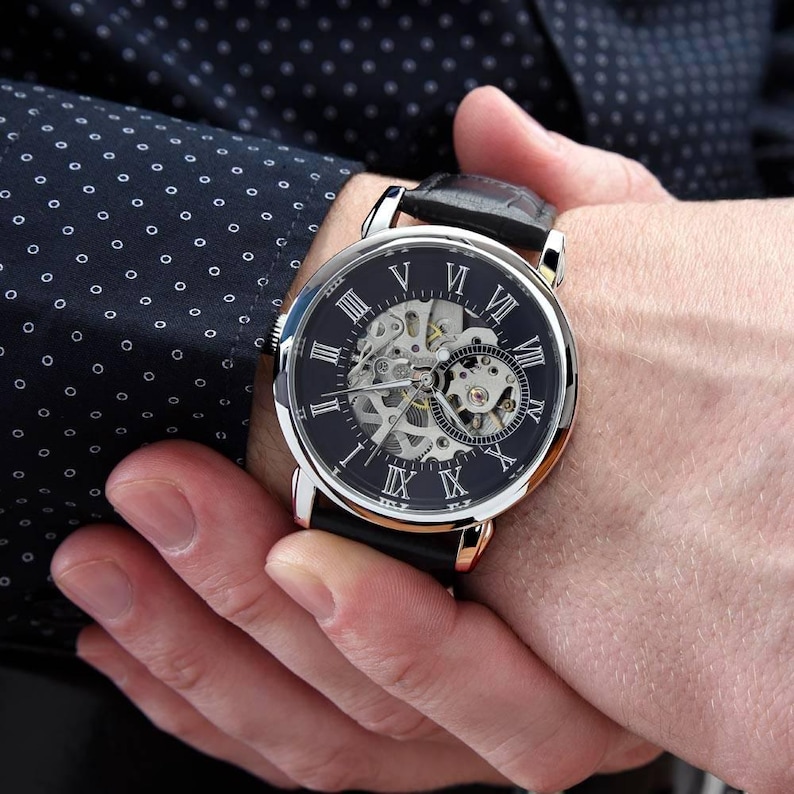 K&ouml;nnte beinhalten: Eine Armbanduhr mit rundem, silberfarbenem Geh&auml;use und schwarzem Zifferblatt mit wei&szlig;en r&ouml;mischen Ziffern. Die Mitte des Zifferblatts ist skelettiert und gibt den Blick auf das mechanische Uhrwerk frei. Die Uhr hat ein schwarzes Lederarmband.
