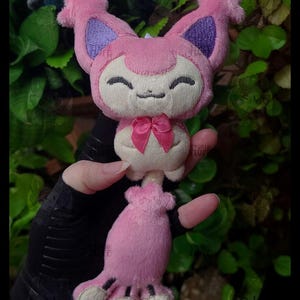May include: A small pink plush toy featuring a cat-like creature with large pointed ears, purple inner ear details, and a smiling expression. The plush has a cream-coloured face and belly, a small pink satin bow at the neck, and a long tail with dark stripes.