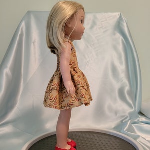 Puede incluir: Una mu&ntilde;eca con un vestido marr&oacute;n y naranja con estampado de hojas y zapatos rojos. La mu&ntilde;eca tiene el pelo rubio y est&aacute; de pie sobre una plataforma gris.