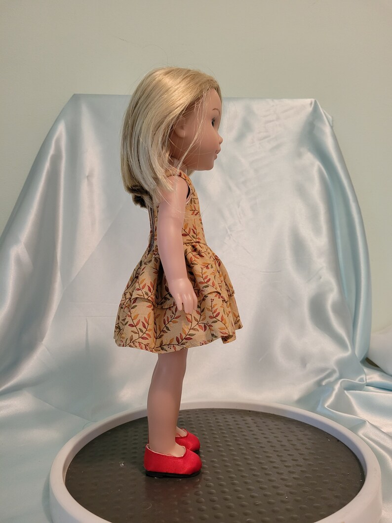 Puede incluir: Una mu&ntilde;eca con un vestido marr&oacute;n y naranja con estampado de hojas y zapatos rojos. La mu&ntilde;eca tiene el pelo rubio y est&aacute; de pie sobre una plataforma gris.