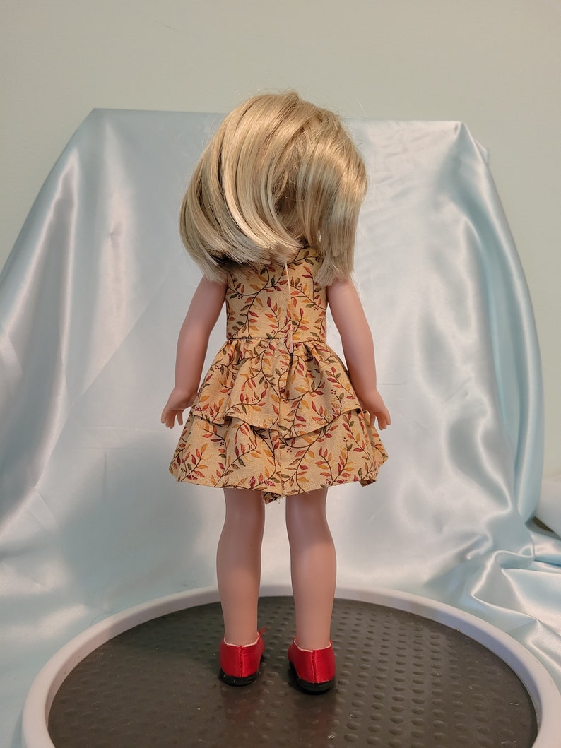 Puede incluir: Una mu&ntilde;eca con un vestido marr&oacute;n y amarillo con estampado de hojas. El vestido tiene una falda con volantes. La mu&ntilde;eca lleva zapatos rojos.