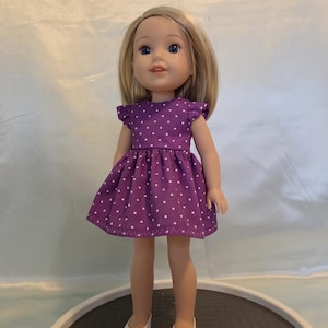 May include: A doll wearing a purple dress with white polka dots and white shoes. The dress has short sleeves with ruffled edges. The doll has blonde hair and blue eyes. The doll is standing on a dark gray surface.