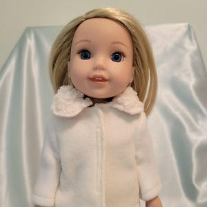 May include: A doll with blonde hair and blue eyes wearing a white coat with a fluffy collar. The doll is also wearing dark blue jeans. The coat is buttoned up and has a soft texture.