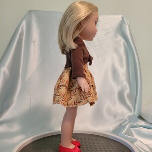 Puede incluir: Una mu&ntilde;eca con un su&eacute;ter marr&oacute;n y un vestido beige con estampado de hojas. La mu&ntilde;eca tiene el pelo rubio y lleva zapatos rojos.