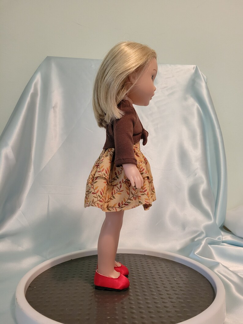 Puede incluir: Una mu&ntilde;eca con un su&eacute;ter marr&oacute;n y un vestido beige con estampado de hojas. La mu&ntilde;eca tiene el pelo rubio y lleva zapatos rojos.