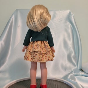 Puede incluir: Una mu&ntilde;eca con un su&eacute;ter verde y una falda marr&oacute;n y amarilla con un estampado de hojas. La mu&ntilde;eca tiene el pelo rubio y lleva zapatos rojos.