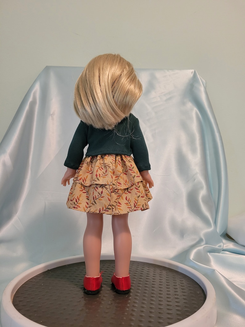 Puede incluir: Una mu&ntilde;eca con un su&eacute;ter verde y una falda marr&oacute;n y amarilla con un estampado de hojas. La mu&ntilde;eca tiene el pelo rubio y lleva zapatos rojos.
