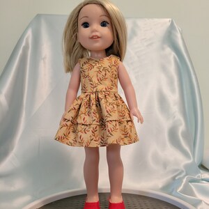 Puede incluir: Una mu&ntilde;eca con un vestido amarillo con un estampado de hojas marrones y rojas. La mu&ntilde;eca tiene el pelo rubio y los ojos azules. La mu&ntilde;eca lleva zapatos rojos.