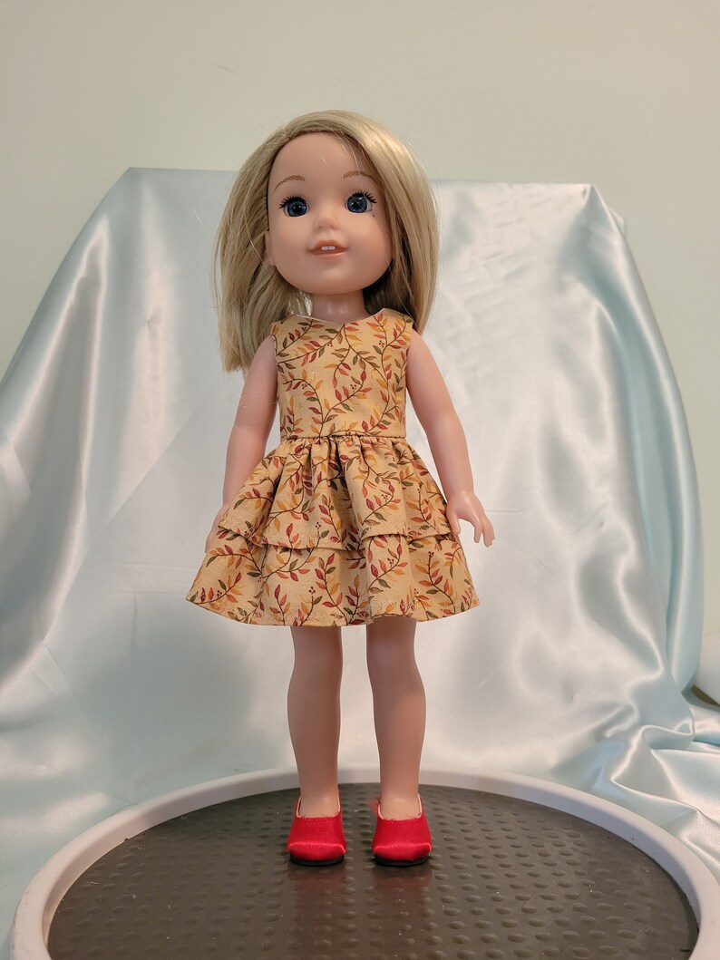 Puede incluir: Una mu&ntilde;eca con un vestido amarillo con un estampado de hojas marrones y rojas. La mu&ntilde;eca tiene el pelo rubio y los ojos azules. La mu&ntilde;eca lleva zapatos rojos.