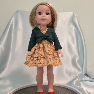 Puede incluir: Una mu&ntilde;eca con un c&aacute;rdigan verde sobre un vestido amarillo con estampado de hojas. La mu&ntilde;eca tiene el pelo rubio y los ojos azules y lleva zapatos rojos.