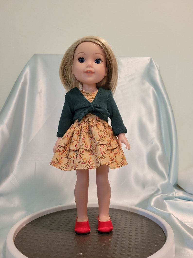 Puede incluir: Una mu&ntilde;eca con un c&aacute;rdigan verde sobre un vestido amarillo con estampado de hojas. La mu&ntilde;eca tiene el pelo rubio y los ojos azules y lleva zapatos rojos.