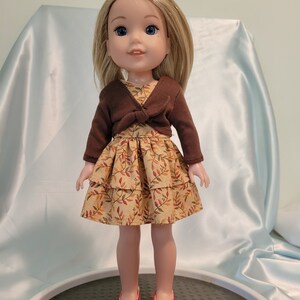 Puede incluir: Una mu&ntilde;eca con un c&aacute;rdigan marr&oacute;n y un vestido de capas con estampado de hojas de oto&ntilde;o. La mu&ntilde;eca tiene el pelo rubio y los ojos azules. Lleva zapatos rojos.