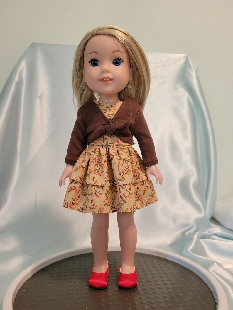Puede incluir: Una mu&ntilde;eca con un c&aacute;rdigan marr&oacute;n y un vestido de capas con estampado de hojas de oto&ntilde;o. La mu&ntilde;eca tiene el pelo rubio y los ojos azules. Lleva zapatos rojos.