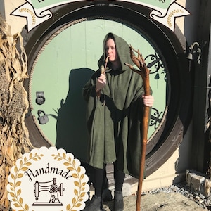 May include: A hooded olive green cloak worn over black leggings and black boots. The outfit includes a long wooden staff with a branch-like top and a pipe held to the mouth. Text at the top reads Woman-Owned Small Business and Handmade.