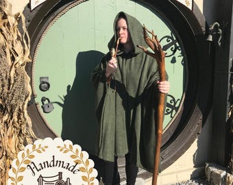 May include: A hooded olive green cloak worn over black leggings and black boots. The outfit includes a long wooden staff with a branch-like top and a pipe held to the mouth. Text at the top reads Woman-Owned Small Business and Handmade.