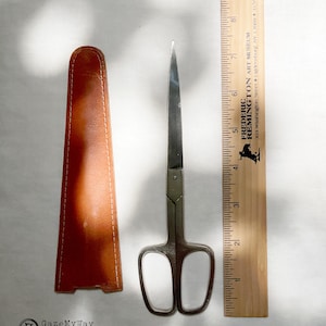 May include: A pair of silver-toned metal scissors with a tan leather sheath, positioned next to a wooden ruler that shows the scissors measure approximately 7.5 inches in length. The ruler features the text Frederic Remington Art Museum.