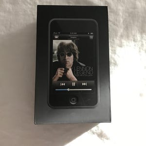 May include: A black iPod device with the Lennon Legend album cover displayed on the screen. The device is in a black box. The screen shows a photo of John Lennon and the word "Imagine".