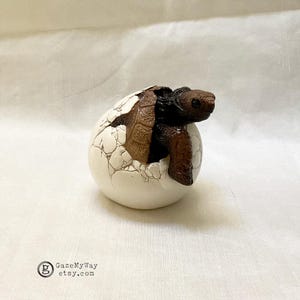 May include: A brown and black turtle hatching from a white egg with a cracked texture. The turtle's head and front legs are visible, emerging from the egg. The egg is on a white surface. The Etsy shop name "GazeMyWay" is visible in the bottom left corner.