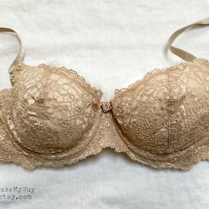 May include: A beige lace bra with adjustable straps. The bra features intricate floral lace detailing on the cups and along the edges. The underwire bra has a small decorative bow between the cups.
