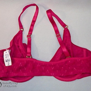 May include: A bright fuchsia bra featuring adjustable shoulder straps with gold-toned hardware and a repeating V-shaped pattern across the band. A white product tag with a barcode and the text 38C is attached to the left side of the garment.