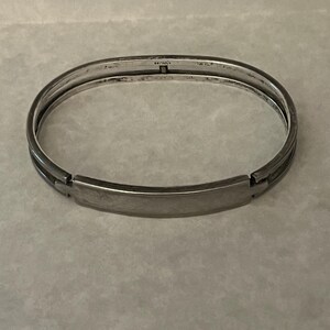 May include: A silver-toned hinged bangle bracelet. The bracelet has a rectangular plate in the center and is marked "STERLING". The bracelet is made of metal and has a simple, elegant design.