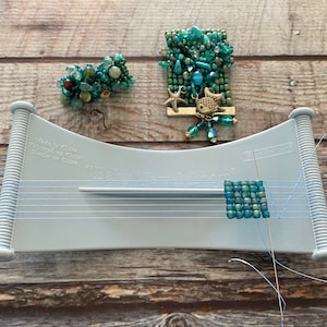 May include: A blue and green beaded bracelet with a silver starfish and fish charm is being made on a blue bead loom. The loom has the text "Easy  Tame  Loom" printed on it.