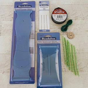 May include: A collection of jewelry-making supplies, including two blue Beadalon Jewel Looms, needles, green WildFire beading thread, green beads, and a wooden button with a starburst design. The items are arranged on a light-colored wooden surface.