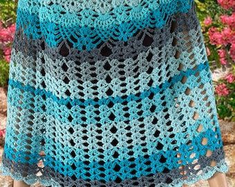 Puede incluir: Un poncho de ganchillo en tonos azules y grises. El diseño de tejido abierto presenta un borde festoneado y un cuello redondo. El degradado de color pasa del azul claro al gris oscuro.