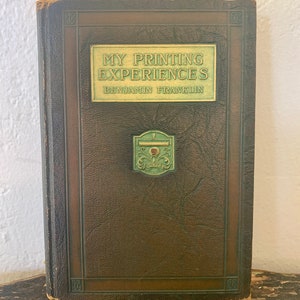 May include: A brown leather-bound book with gold lettering on the spine and front cover. The title is "My Printing Experiences" by Benjamin Franklin. The book has a green and gold decorative design on the front cover.