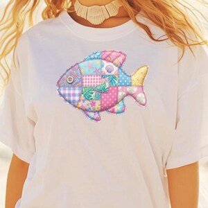 May include: White t-shirt with a vibrant patchwork fish motif. The fish is made up of different patterned fabrics, such as floral, polka dot, and seashell designs. The design is centrally placed on the shirt, giving it a fun and whimsical look.