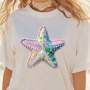 May include: White t-shirt featuring a colorful, patchwork starfish design. The starfish is made of various patterned fabrics, including floral, gingham, and seashell prints. The edges are stitched with a contrasting thread.