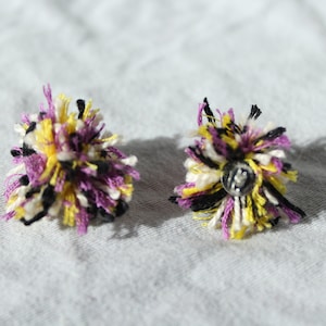 Könnte beinhalten: Ein Paar Ohrringe mit Bommeln aus schwarzem, weißem, gelbem und violettem Garn. Die Bommeln sind an silberfarbenen Metallsteckern befestigt.