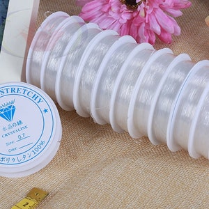 May include: A close-up of a roll of clear elastic string for jewelry making. The string is wound around a white spool with a blue label that reads "Strong & Stretchy" and "Crystaline 0.7".