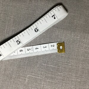May include: A white tape measure with black numbers, showing measurements from 2 to 7 inches. The tape measure is folded in half and the end is secured with a gold metal clasp.