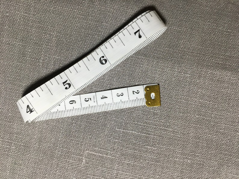May include: A white tape measure with black numbers, showing measurements from 2 to 7 inches. The tape measure is folded in half and the end is secured with a gold metal clasp.
