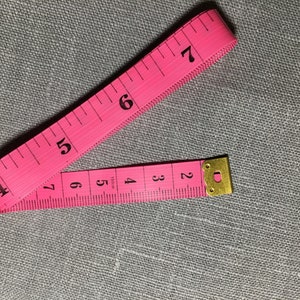 May include: A pink measuring tape with black numbers from 2 to 7. The tape is folded in half and the end is secured with a gold metal clasp.