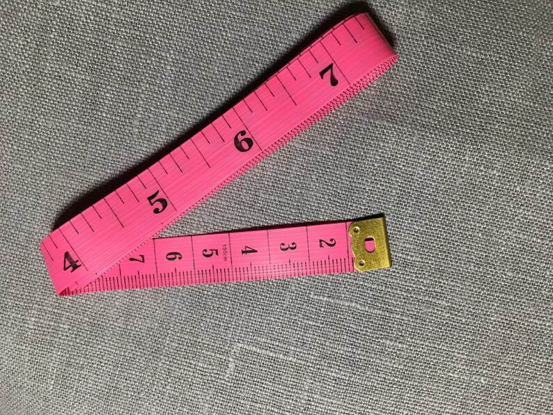 May include: A pink measuring tape with black numbers from 2 to 7. The tape is folded in half and the end is secured with a gold metal clasp.