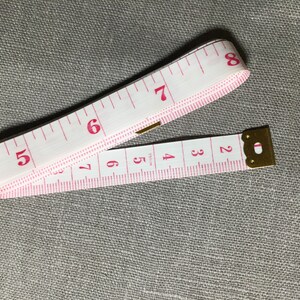 May include: A white measuring tape with pink markings and a gold metal end. The tape is extended to show numbers 1 through 8.